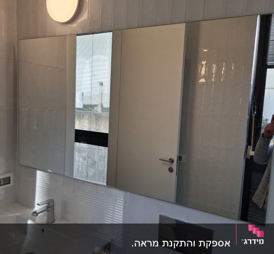 מראה רחבה מותקנת על קיר אריחים בחדר רחצה מעל הכיור